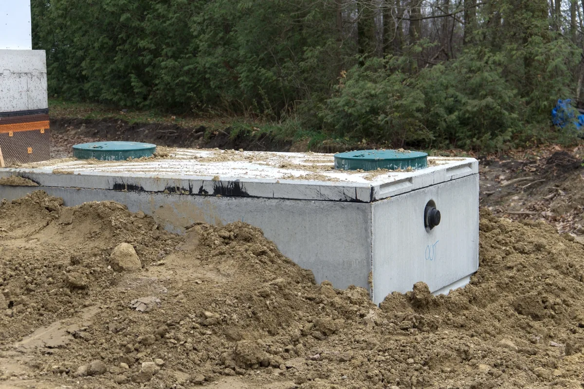 a large, sturdy concrete septic tank being carefully installed in the ground, surrounded by a team of workers considering the optimal placement for efficient wastewater management. A residential septic tank design features a concrete structure partially buried in soil, with two green access covers prominently visible on top. Surrounded by lush trees and vegetation, it seamlessly blends into the natural landscape.