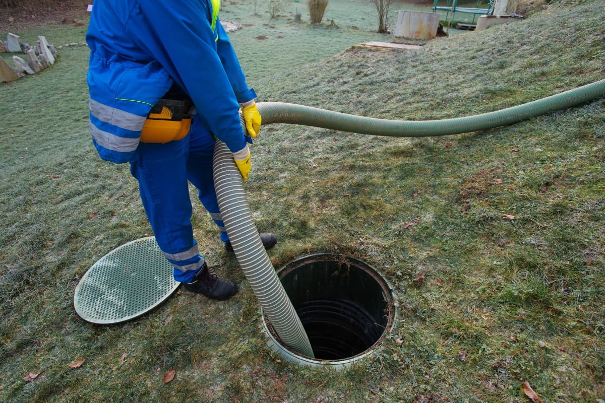 A worker in blue overalls handles a hose connected to an open septic tank on a grassy area, swiftly addressing potential septic emergencies.