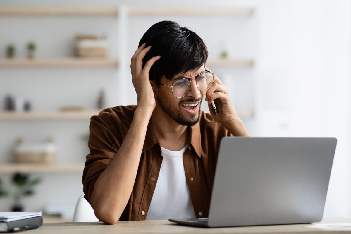 A man with glasses, looking frustrated, talks on the phone about septic emergencies while using a laptop at a desk.