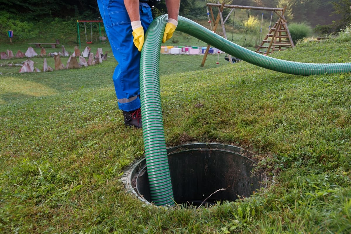 A worker in blue overalls and yellow gloves inserts a green hose into an open septic tank on grassy terrain near a playground, conducting a routine septic system inspection.