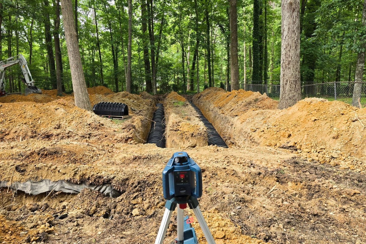 A construction laser level overlooks dug trenches with drainage pipes in a forested area, surrounded by piles of dirt and trees, preparing the groundwork for septic system leach fields.