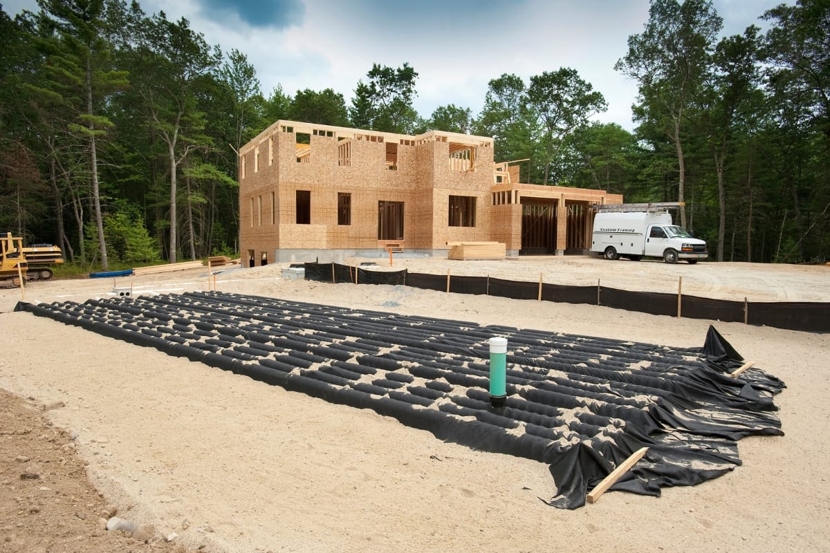 A two-story house under construction with wooden frames, surrounded by trees, awaits its septic system installation. A white van is parked nearby, and construction materials are laid out in the foreground, alongside newly marked leach fields.