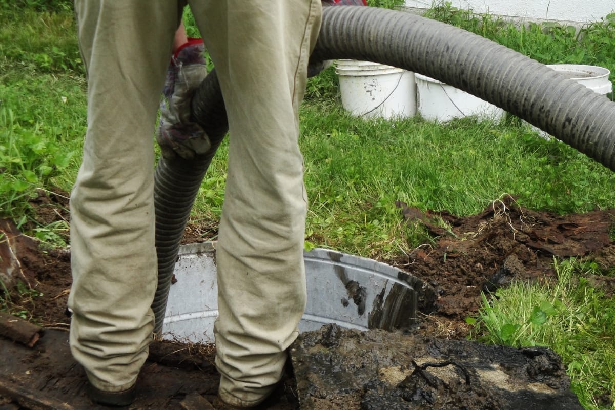 A person is holding a hose while standing near an open septic tank in a grassy area, ensuring the smooth operation of the septic system. White buckets are visible in the background, possibly used for managing leach fields nearby.