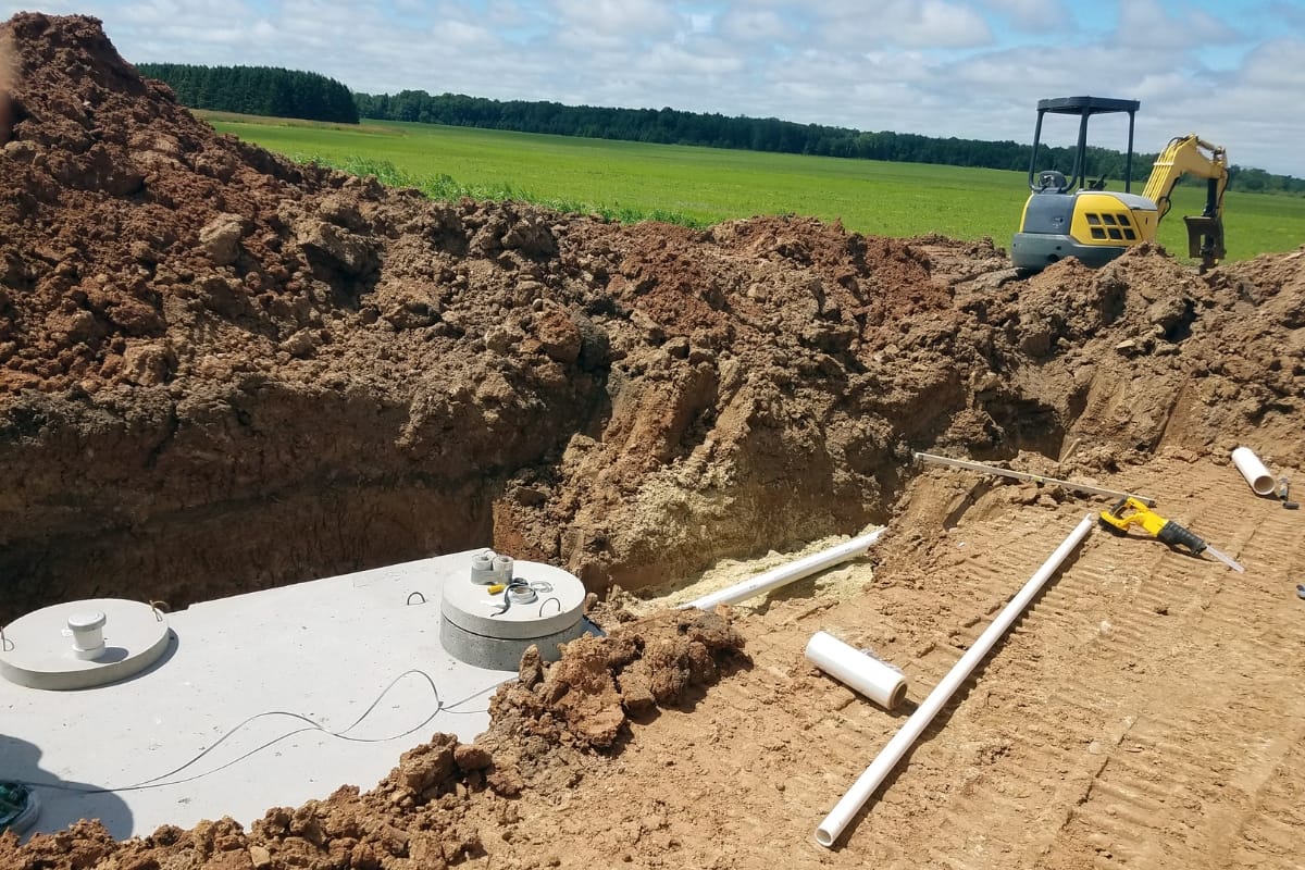 A construction site showcases an excavated pit with a concrete septic tank being expertly installed as part of the new septic system. A small excavator and tools are nearby, while fields and leach fields stretch towards a backdrop of trees.