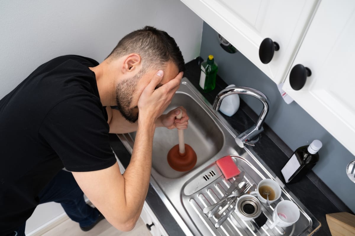 A person tackling a clogged kitchen sink with a plunger, surrounded by unwashed dishes and cleaning supplies, wonders if septic tank issues are to blame for the persistent plumbing woes.