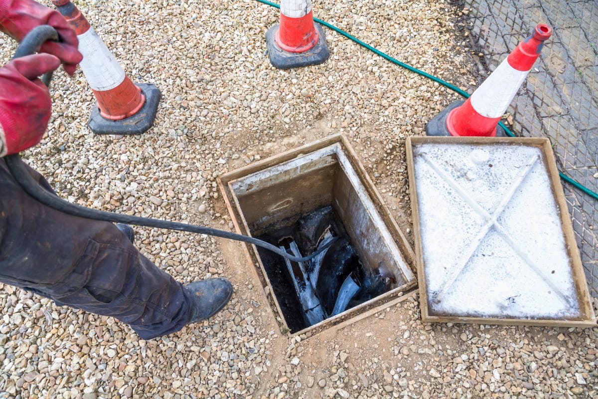 A person wearing red gloves is standing next to an open utility box with cables inside, perhaps gathering insights akin to septic tank maintenance tips. Orange traffic cones are carefully positioned on the gravel surface, marking the area with precision.