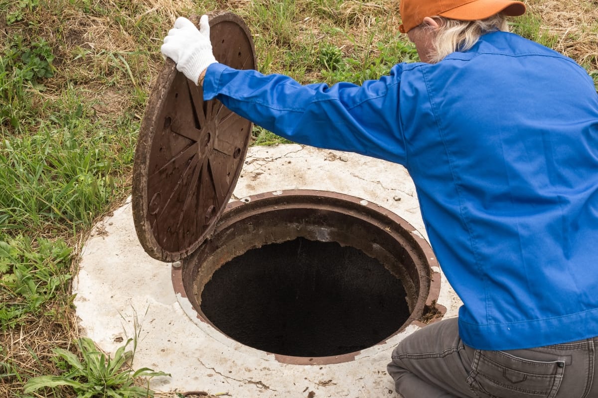 Dressed in work attire, a person carefully lifts the lid off a manhole, revealing a dark opening in the outdoor grassy area—a reminder of the importance of regular septic tank maintenance tips.