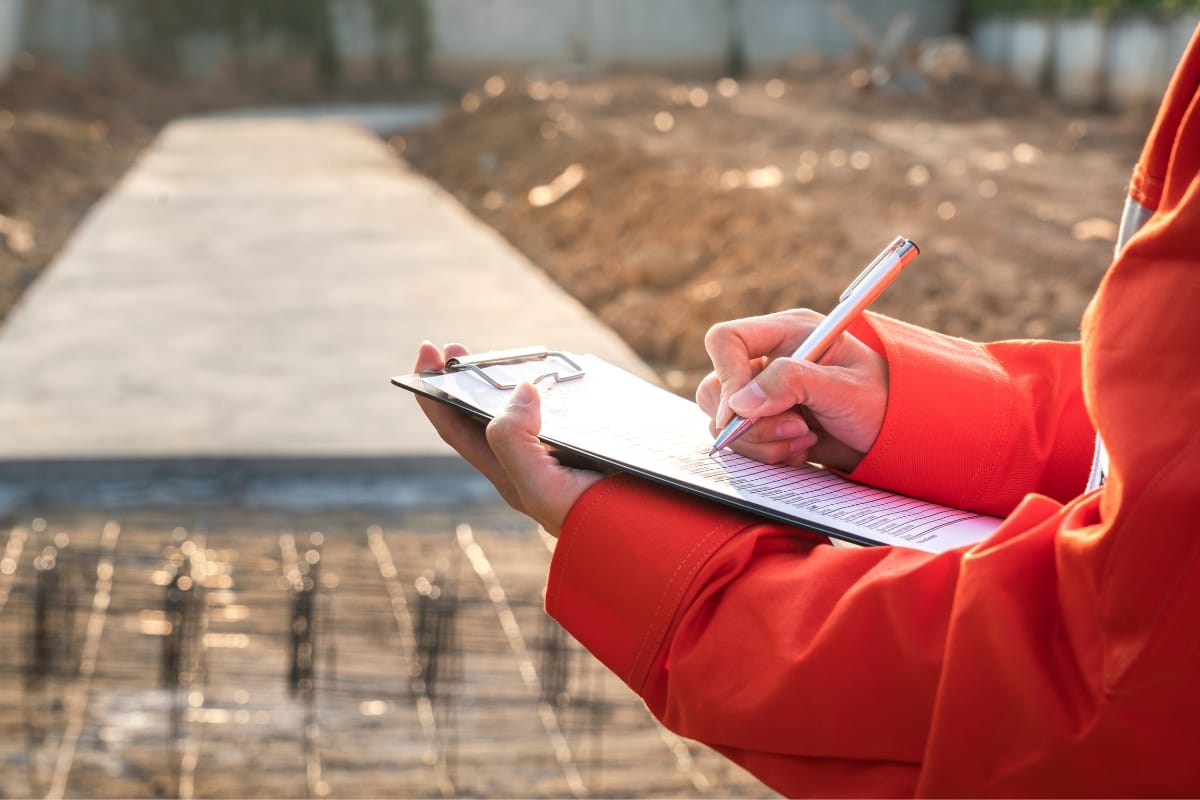 A person in an orange jacket conducts a site assessment, writing diligently on a clipboard at the construction site, surrounded by a visible pathway and soil.