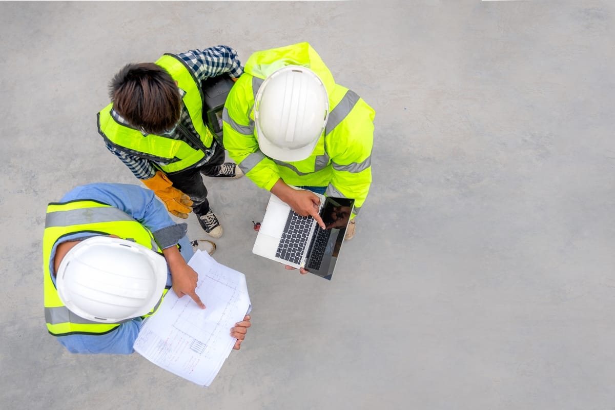 Three construction workers in safety gear conduct a site assessment as they review a blueprint and use a laptop on the concrete surface.