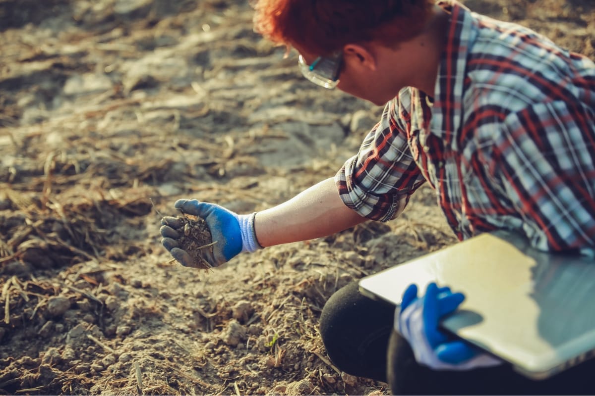 A person with red hair, wearing gloves and safety glasses, conducts a site assessment in a barren field, examining soil while holding a laptop.