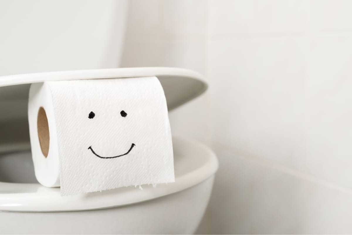 A toilet with a closed lid hosts a roll of toilet paper, noted for being the best toilet paper for septic tanks, playfully adorned with a smiling face drawn in black marker. The setting is completed by a tiled bathroom wall.