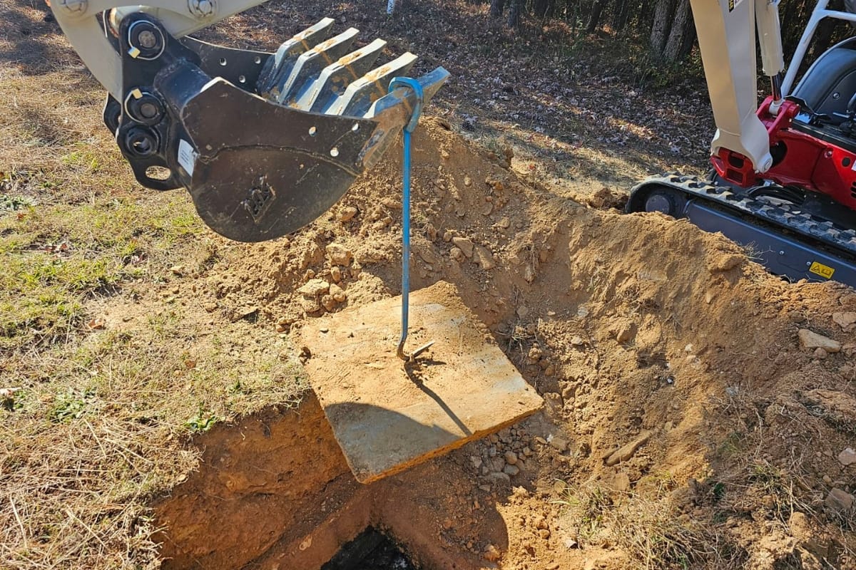 Blog 28 At a dirt excavation site, an excavator bucket effortlessly lifts a concrete slab with a hook. Workers are reminded to also know how to find your septic tank lid during such operations, ensuring smooth progress and safety.