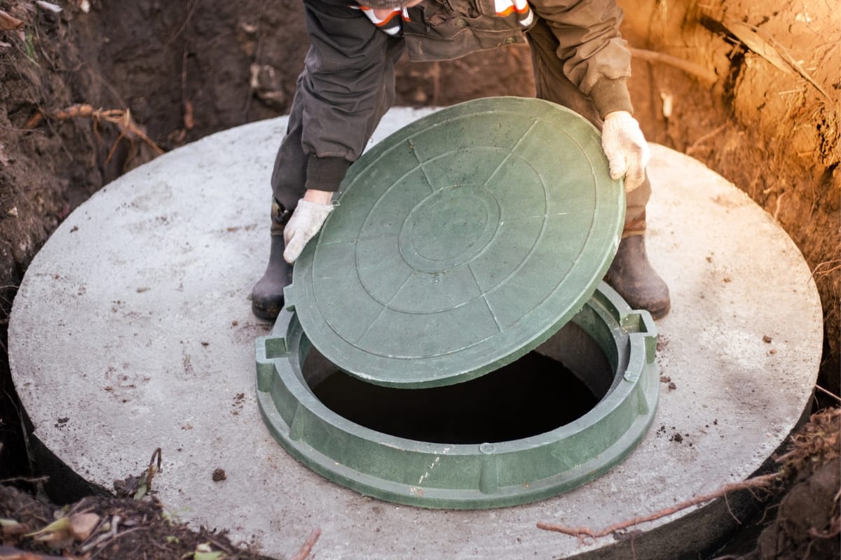 The person carefully lifts the green lid of a concrete septic tank, inspecting it for potential issues. They're focused on septic tank alarm troubleshooting, ready to dive into identifying causes and quick fixes.