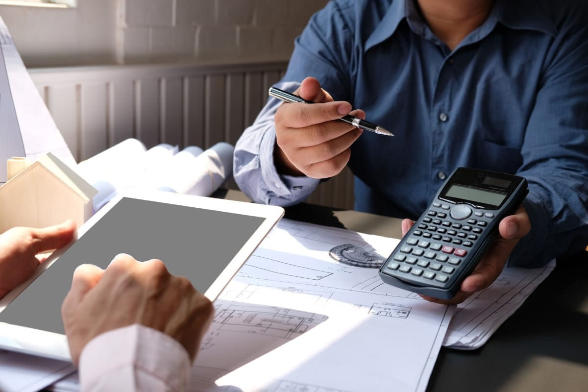 Two people discuss architectural plans on a table with a tablet, calculator, and pen, carefully considering every detail, including the septic tank cost, to ensure the project stays within budget.