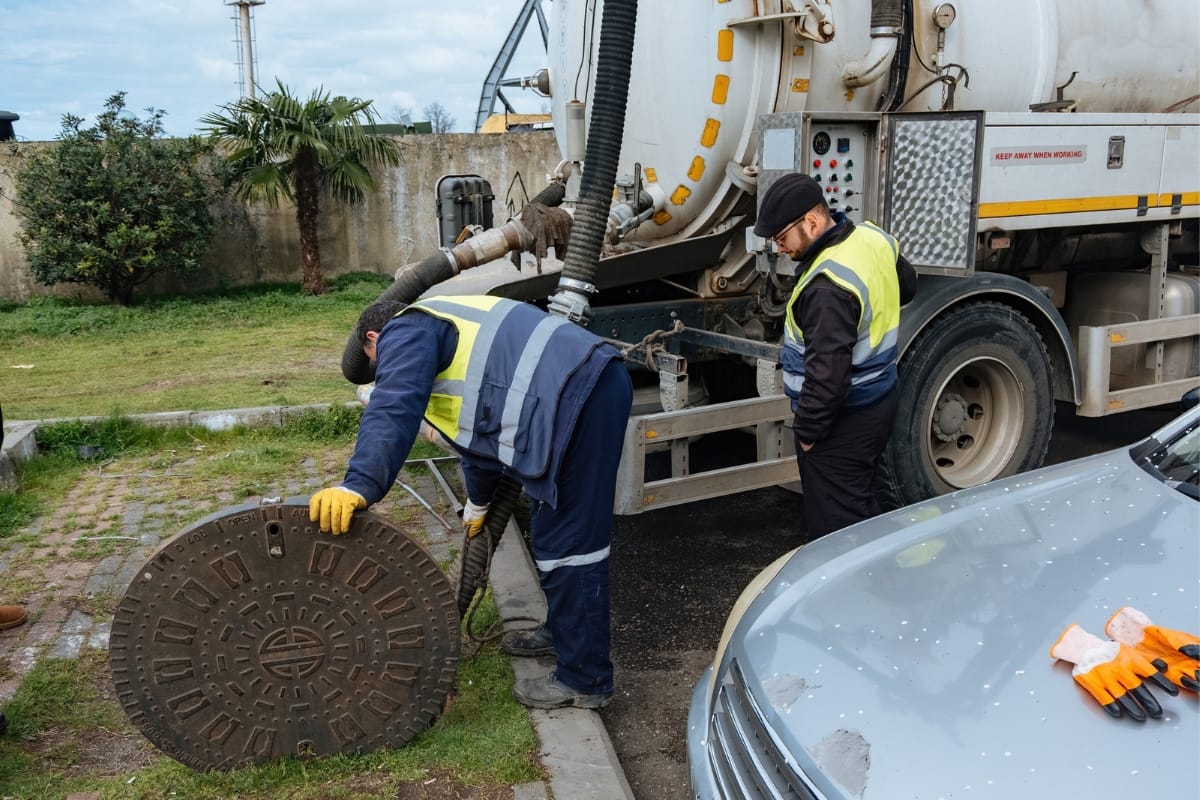 Two workers in safety gear are lifting a manhole cover next to a vacuum truck, addressing why the septic tank keeps filling with water. They're on a paved area with grass and a parked car nearby.