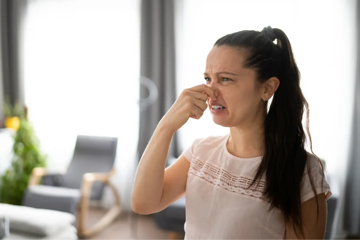 Warning Signs Your Septic Tank Needs Pumping 2 A woman in a light pink shirt holds her nose with a disgusted expression, standing in a bright room with gray curtains and furniture. It might be one of those signs your septic tank needs pumping.