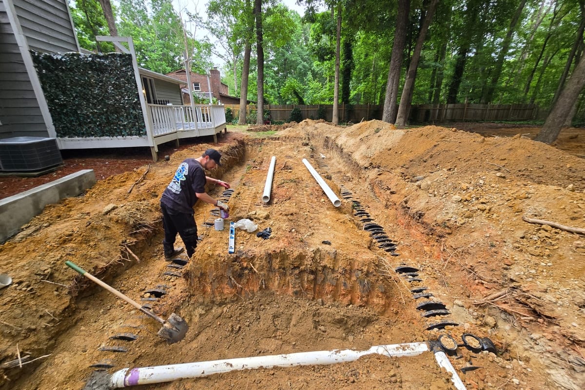 A person expertly works on drainage pipes in a residential backyard, strategically placing them in dug trenches amidst nearby trees, ensuring equipment longevity throughout the installation process.