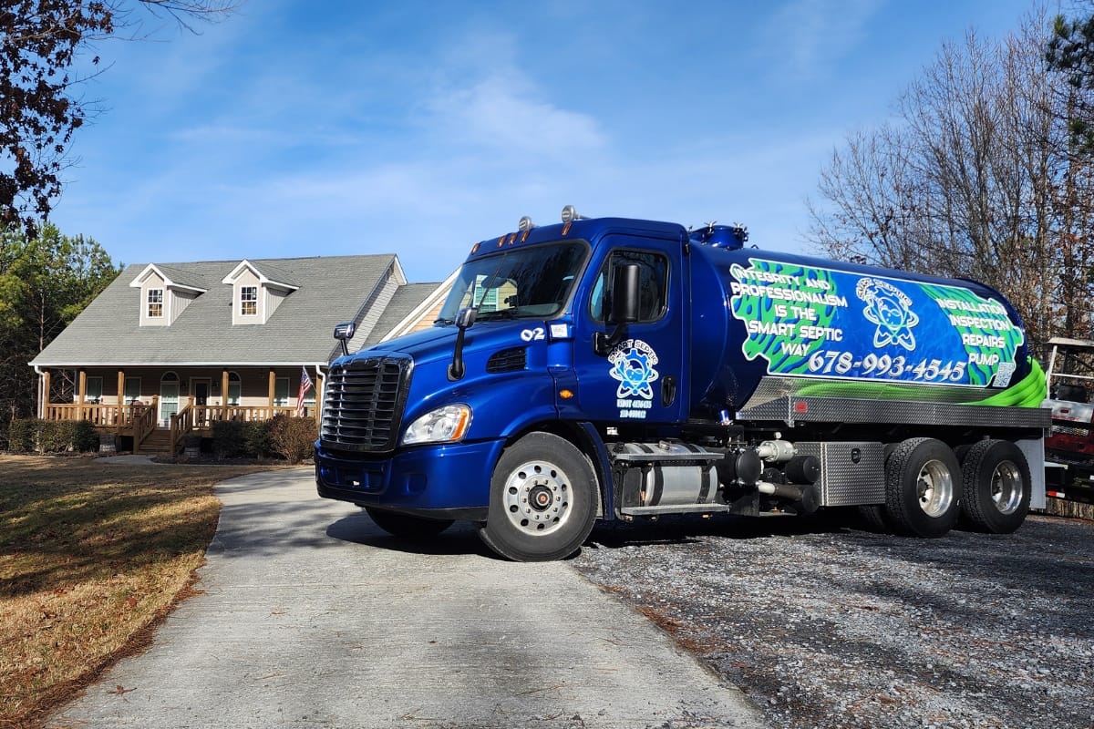 DIY Septic System Maintenance Georgia: Essential Tips for Homeowners 7 A blue septic service truck, offering Septic Tank Inspections in Canton, is parked on a residential driveway, right in front of a charming house with a porch. The truck proudly displays the company's information and phone number for easy contact.
