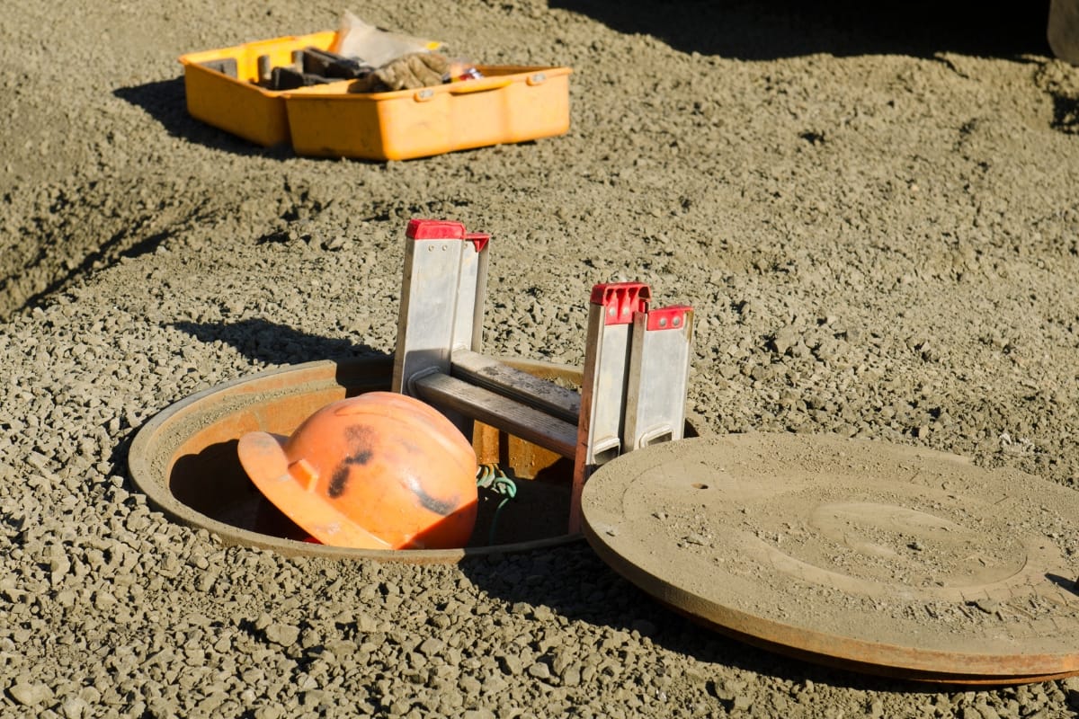 Buying a Home with a Septic System in Acworth, GA: Essential Insights 5 An open manhole with a hard hat and a collapsible ladder inside, surrounded by dirt—and a yellow tool box in the background—could represent some of the inspections involved when buying a home with a septic system.