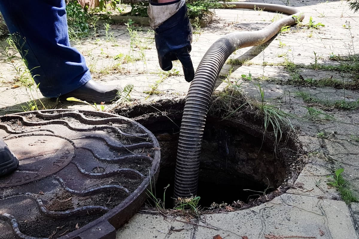 Off-Grid Septic Systems in Marietta, GA: Solutions for Remote Homes 3 Close-up of a person using a hose to clean or pump out a manhole, often part of off-grid septic systems, with the cover removed; paving and vegetation are visible around the opening.
