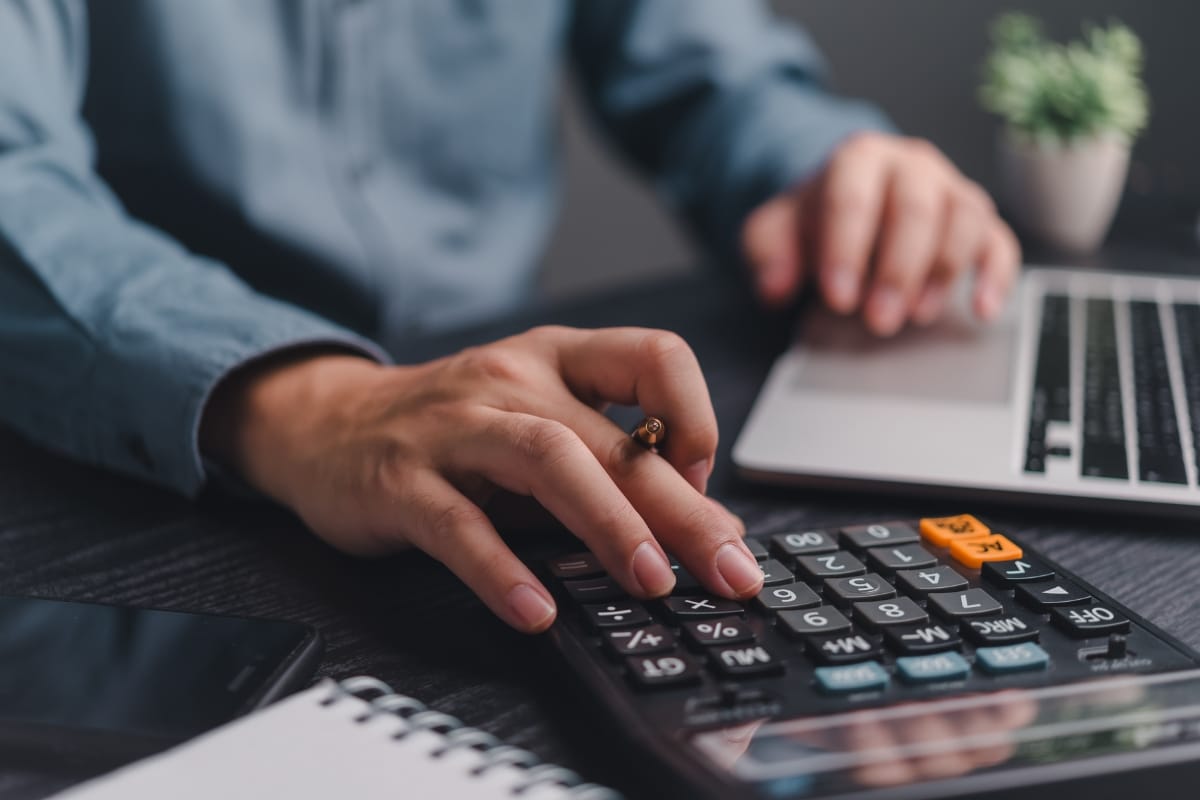 Off-Grid Septic Systems in Marietta, GA: Solutions for Remote Homes 7 A person uses a calculator with one hand and types on a laptop with the other, researching off-grid septic systems. A notebook, pen, and plant are visible on the desk.