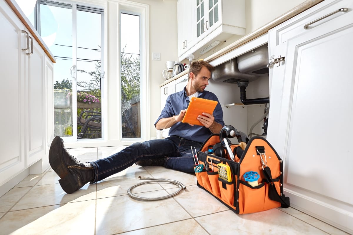 Septic System Summer Care in Woodstock: Preparing for Gatherings 5 A plumber sits on a kitchen floor with tools and equipment, looking at a tablet while inspecting pipes under a sink, ensuring the plumbing connects properly to the home's septic system.