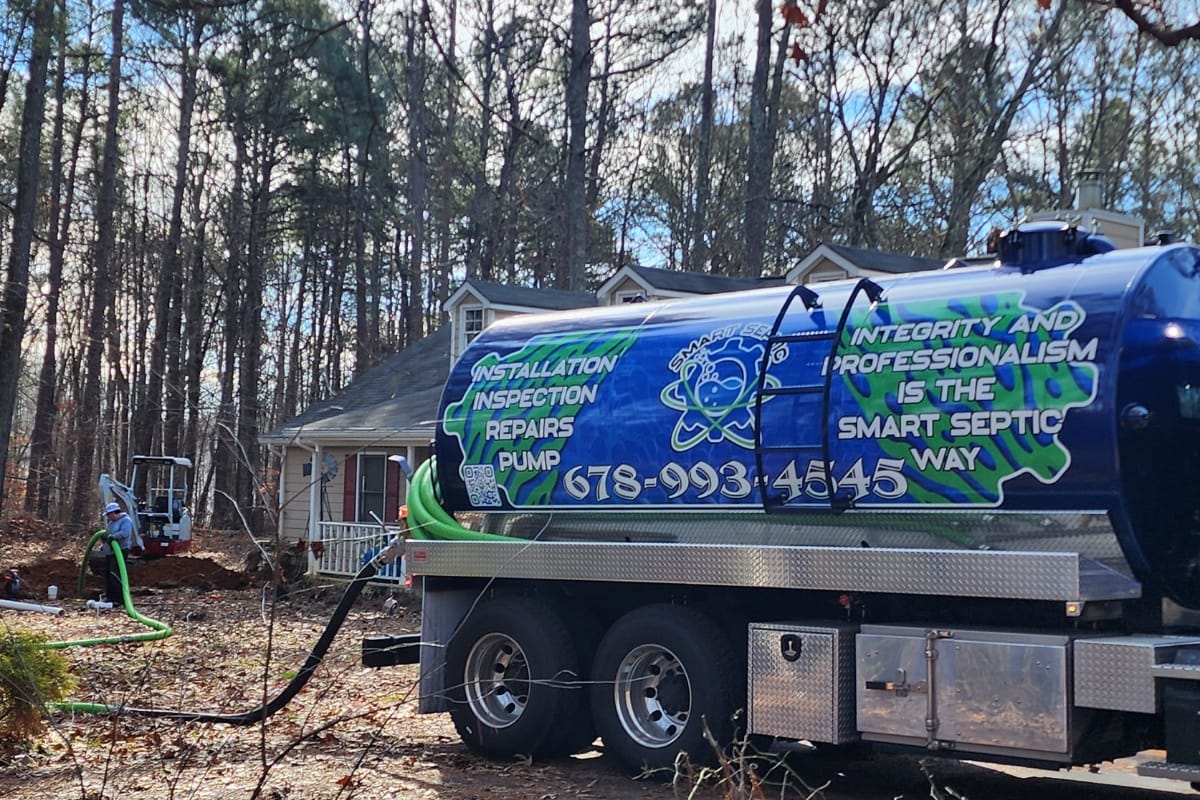 Septic System Failure Causes in Kennesaw, GA: How to Prevent Them 5 A septic service truck is parked in front of a house surrounded by trees, with hoses connected to the home's septic system, addressing a possible septic system failure as workers attend to the issue in the background.