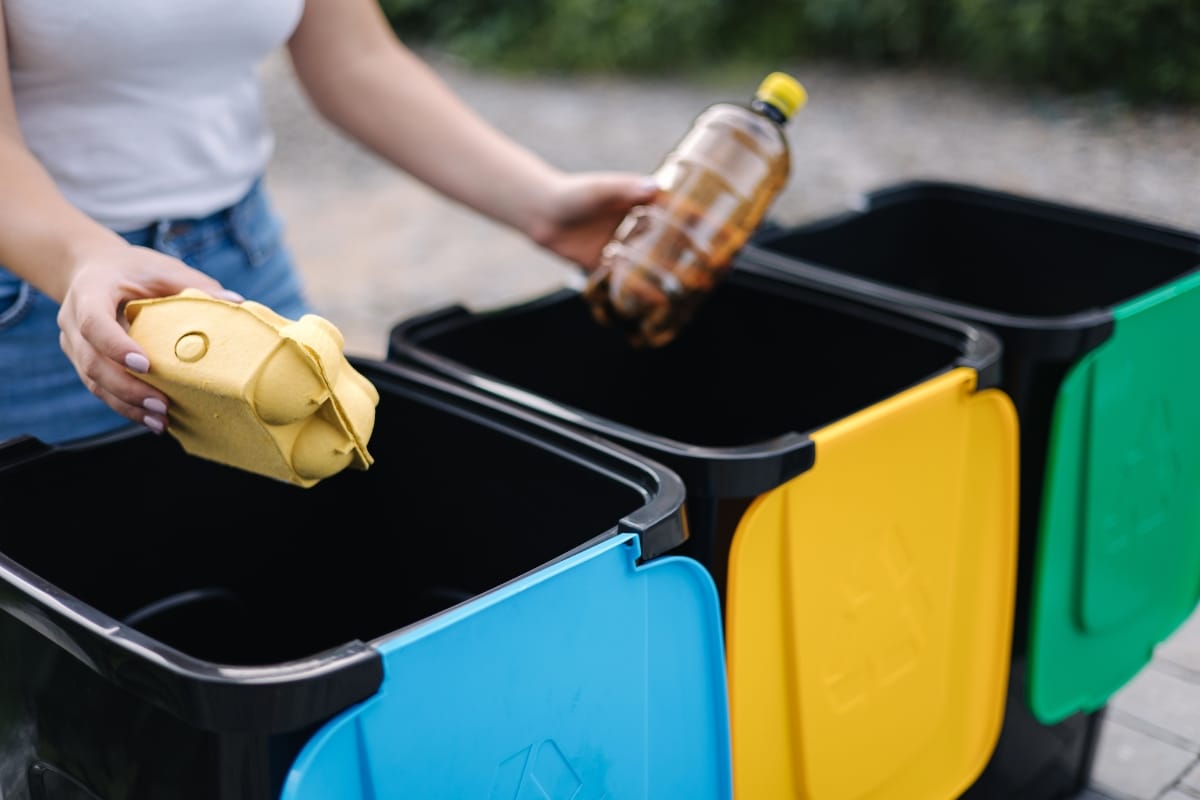 Septic System Failure Causes in Kennesaw, GA: How to Prevent Them 7 Person sorting waste into recycling bins: holding an egg carton over a blue bin and a plastic bottle over a yellow bin, with a green bin also present—smart disposal habits that help prevent issues like septic system failure.