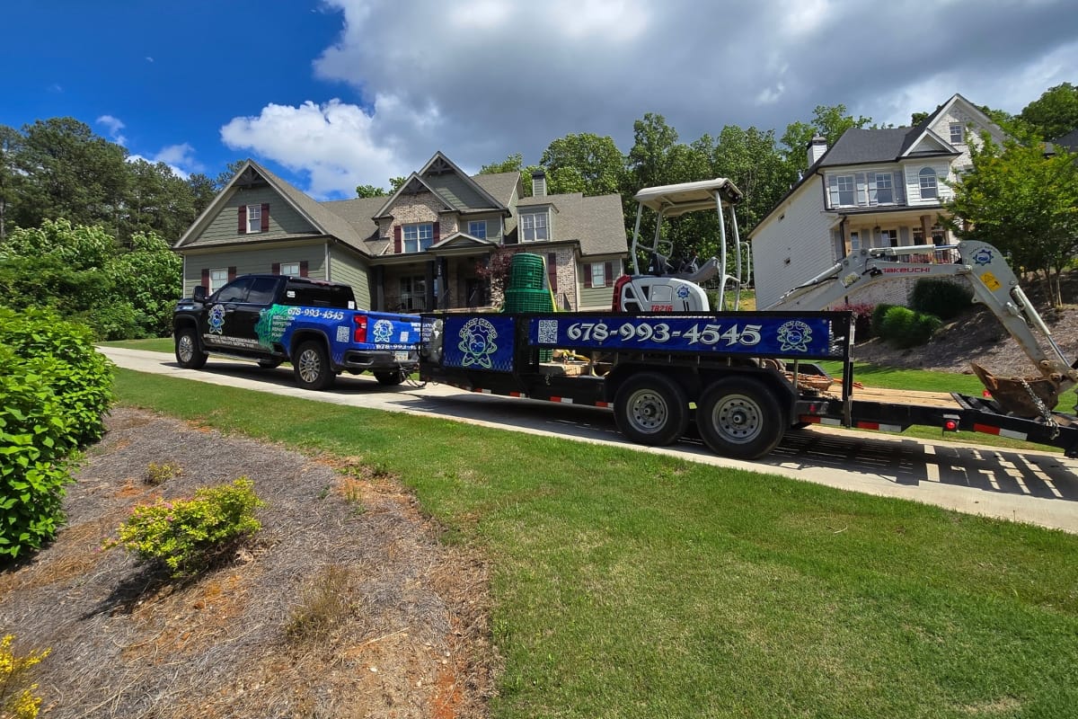 Septic System Inspection Process in Marietta, GA: What to Expect 3 A truck with a trailer carrying landscaping equipment, possibly for a septic system inspection, is parked in front of suburban houses on a sunny day.