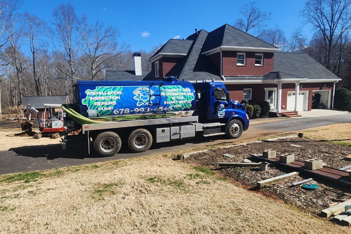 Septic System Inspection Process in Marietta, GA: What to Expect 4 A blue septic service truck is parked on the driveway in front of a two-story brick house with a garage, ready for a septic system inspection on a sunny day.