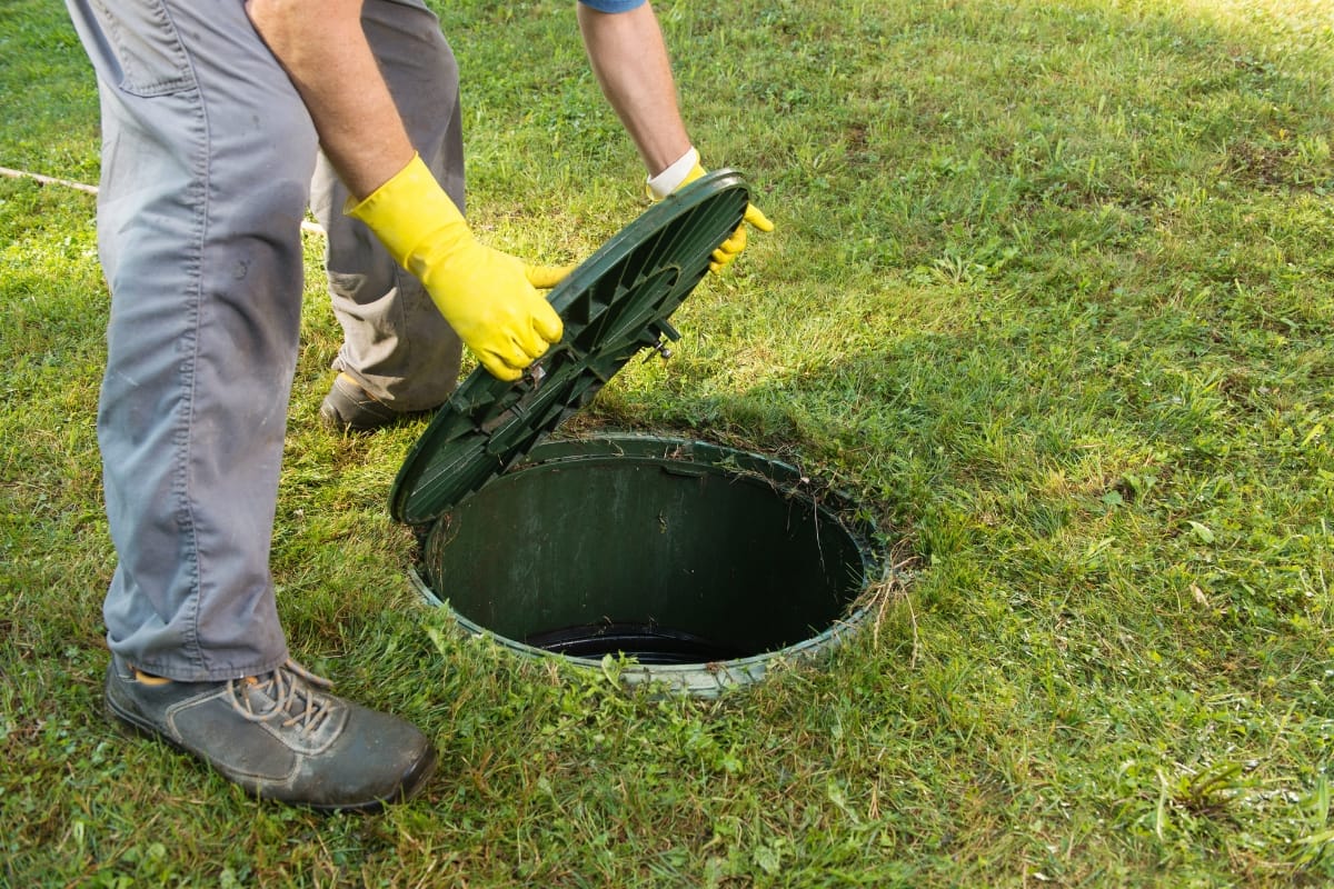 Septic System Inspection Process in Marietta, GA: What to Expect 6 Person wearing gloves lifts the lid of an underground septic tank on a grassy lawn during a septic system inspection.