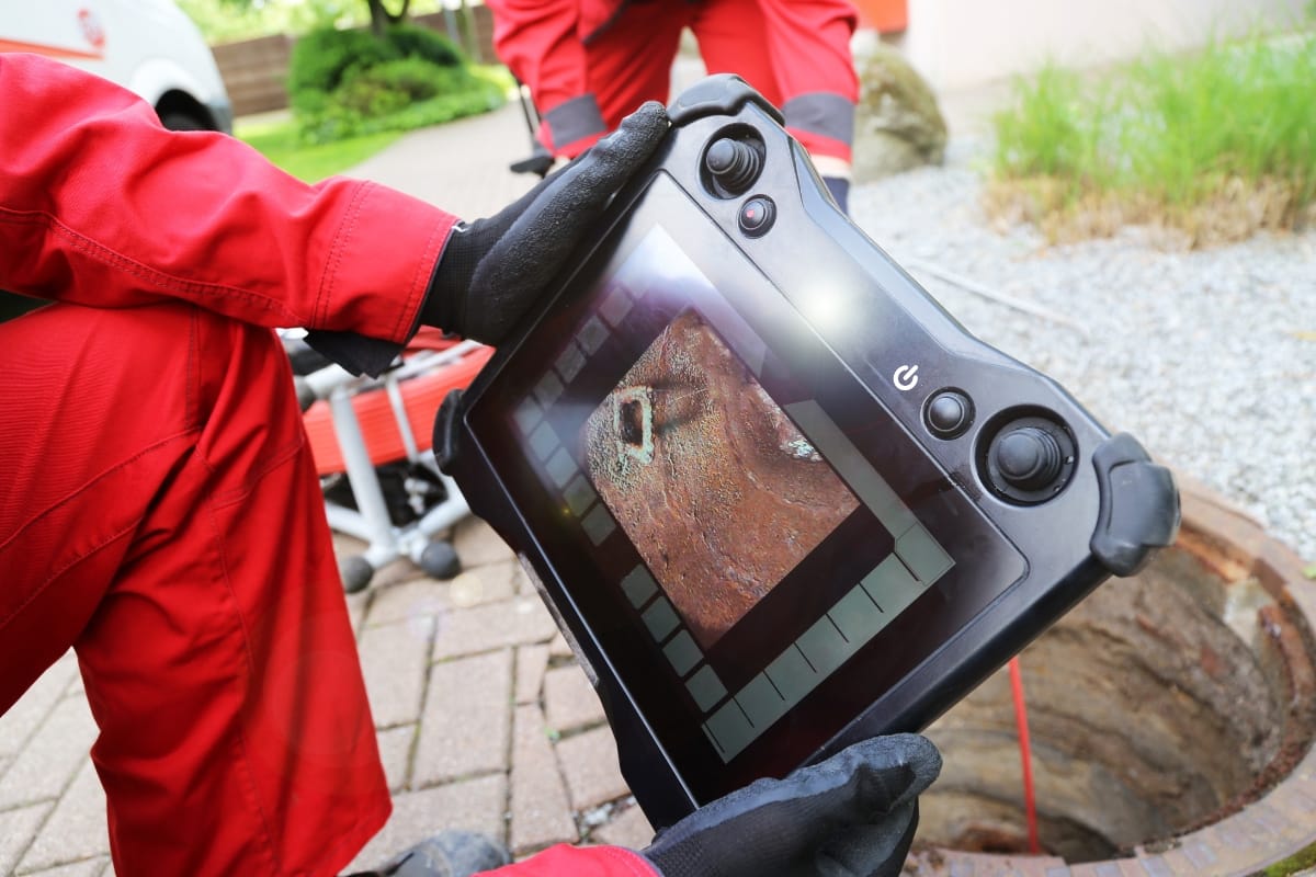 A person in red protective clothing and gloves holds a tablet displaying an image of a rusted object, standing near an open manhole outdoors, likely inspecting or maintaining a septic system.