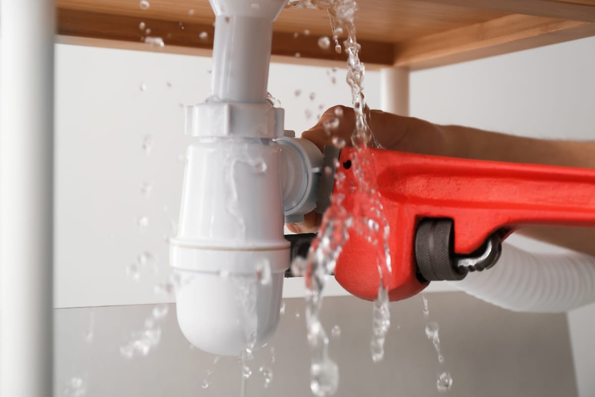 Plumbing and Septic Systems in Woodstock, GA: How They Work Together 10 Water leaking from a white pipe under a sink while a person tightens a fitting with a red pipe wrench, highlighting common plumbing and septic systems issues.