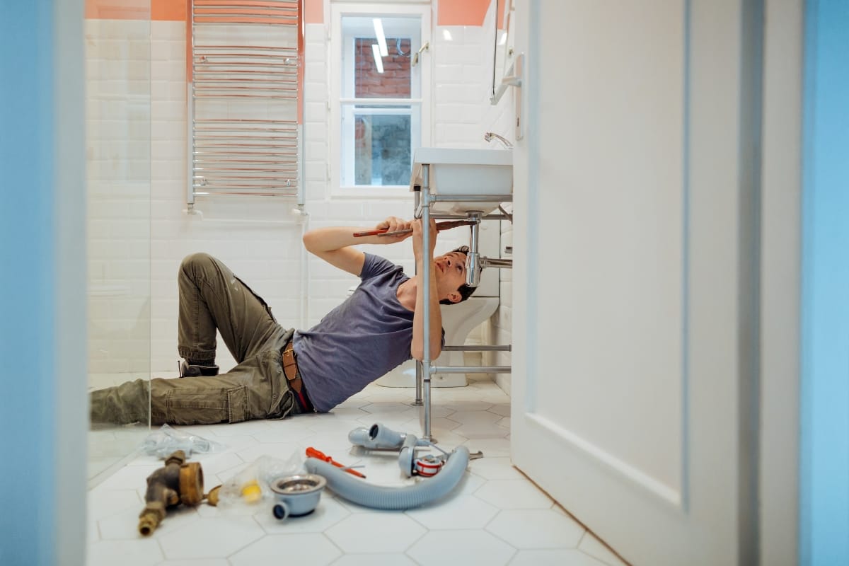 Plumbing and Septic Systems in Woodstock, GA: How They Work Together 11 A person lies on a bathroom floor repairing pipes under a sink, with tools and plumbing parts scattered nearby—clear evidence of hands-on work with plumbing and septic systems.