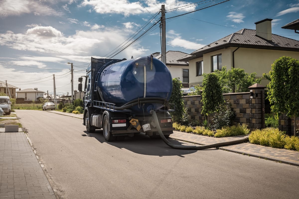 Septic System Myths in Kennesaw, GA: Debunking Common Misconceptions 1 A large blue sewage tanker truck, often associated with Septic System Myths, is parked on a residential street with a hose extended toward a house.
