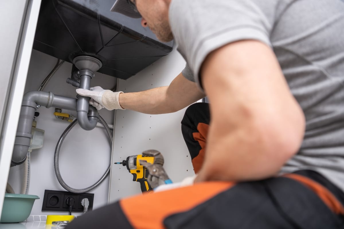 Septic System Myths in Kennesaw, GA: Debunking Common Misconceptions 4 A plumber wearing gloves repairs pipes under a sink, with tools and a drill nearby—dispelling common septic system myths through expert care.