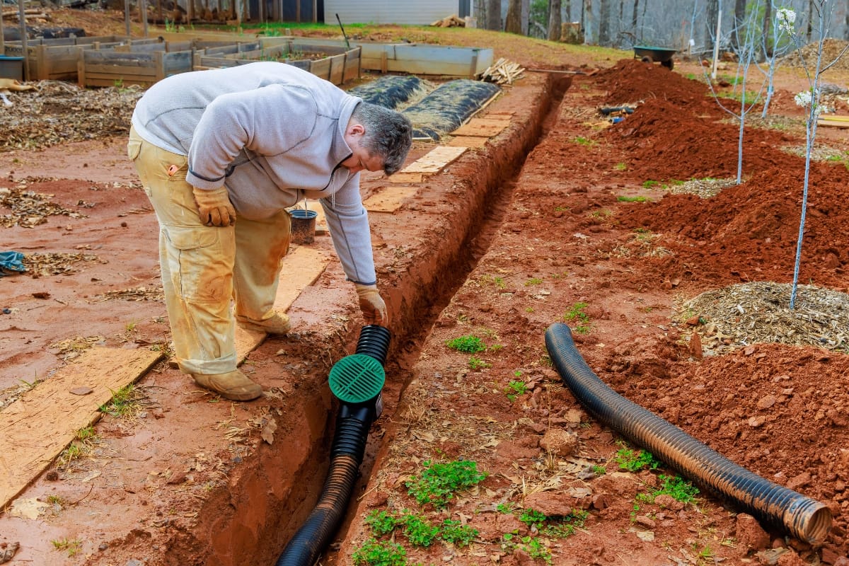 Septic System Myths in Kennesaw, GA: Debunking Common Misconceptions 5 A person installs a black plastic drainage pipe with a green cover in a trench dug through red soil in a garden area, helping to dispel common Septic System Myths about modern drainage solutions.