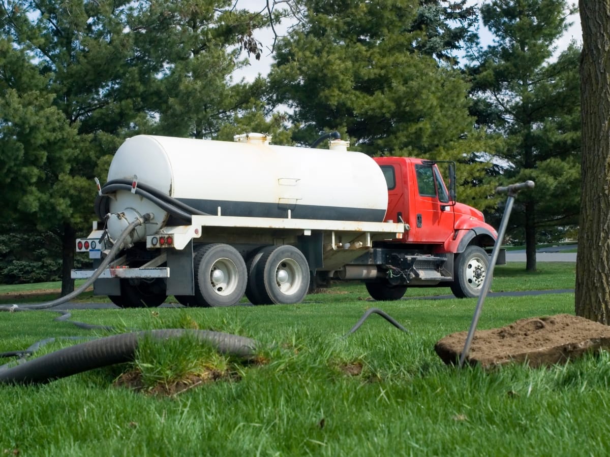 Fall Septic Maintenance in Acworth, GA: Preparing Your System 11 A red and white septic tank truck, used for septic maintenance, is parked on grass with hoses connected and extending into the ground nearby.