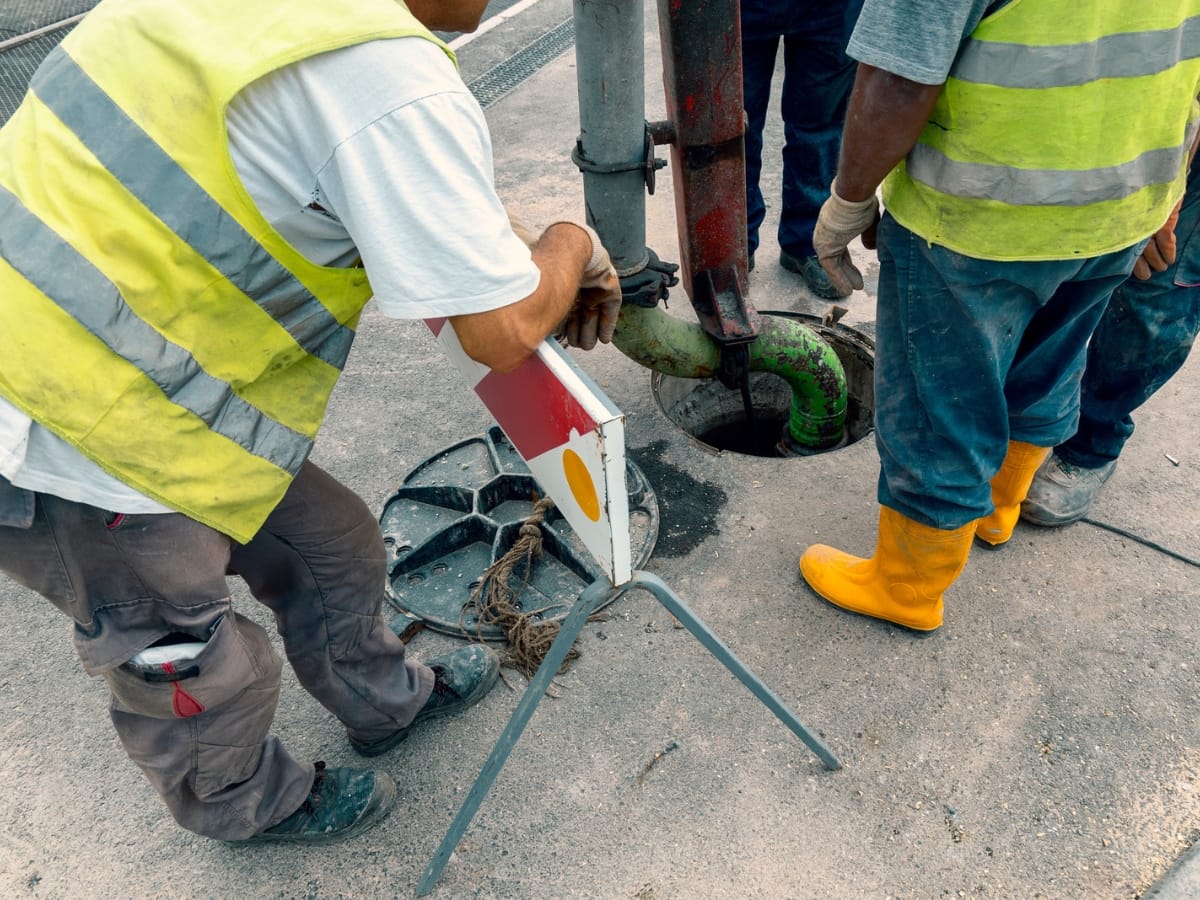 Septic System Components in Marietta, GA: Understanding the Basics 9 Three workers in safety vests operate equipment at an open manhole, inspecting septic system components. One holds a caution sign while another, in yellow boots, ensures safety on the street.