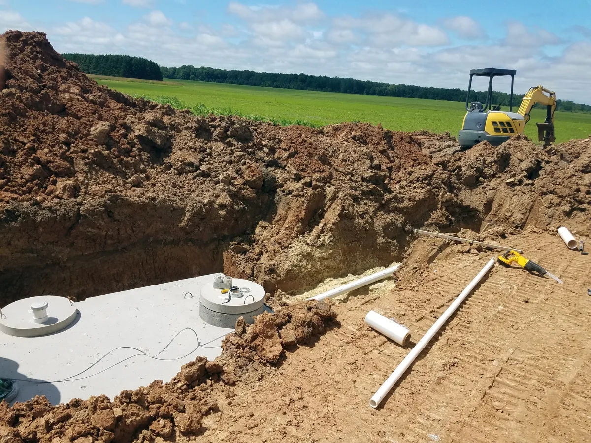 A large trench with a newly installed concrete septic tank, surrounded by dirt piles, PVC pipes, and construction equipment in a rural field, highlights key septic system components in the installation process.