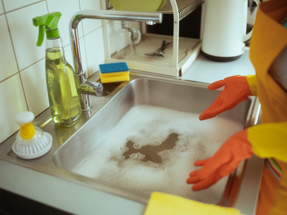 Emergency Septic Services Marietta, GA: When to Call the Pros 1 A person wearing orange gloves stands by a kitchen sink filled with soapy water, cleaning supplies, and sponges nearby—reminding homeowners of the importance of Emergency Septic Services Marietta for maintaining a healthy household.