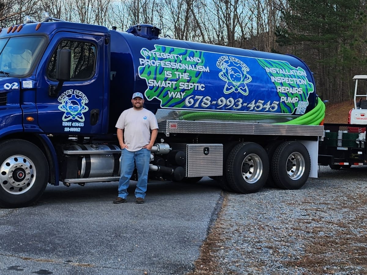 Septic Maintenance Cost in Woodstock, GA: The Price of Neglect 2 A man stands in front of a septic service truck displaying company branding and contact information, parked on a paved lot with trees in the background, highlighting affordable septic maintenance cost options.
