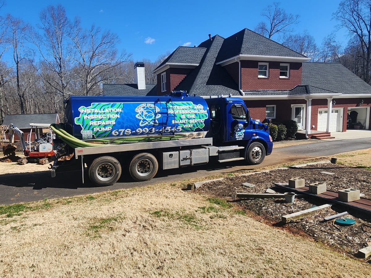 A blue septic service truck with company branding is parked on the driveway of a large brick house on a sunny day, highlighting the importance of regular service to help manage your septic maintenance cost.