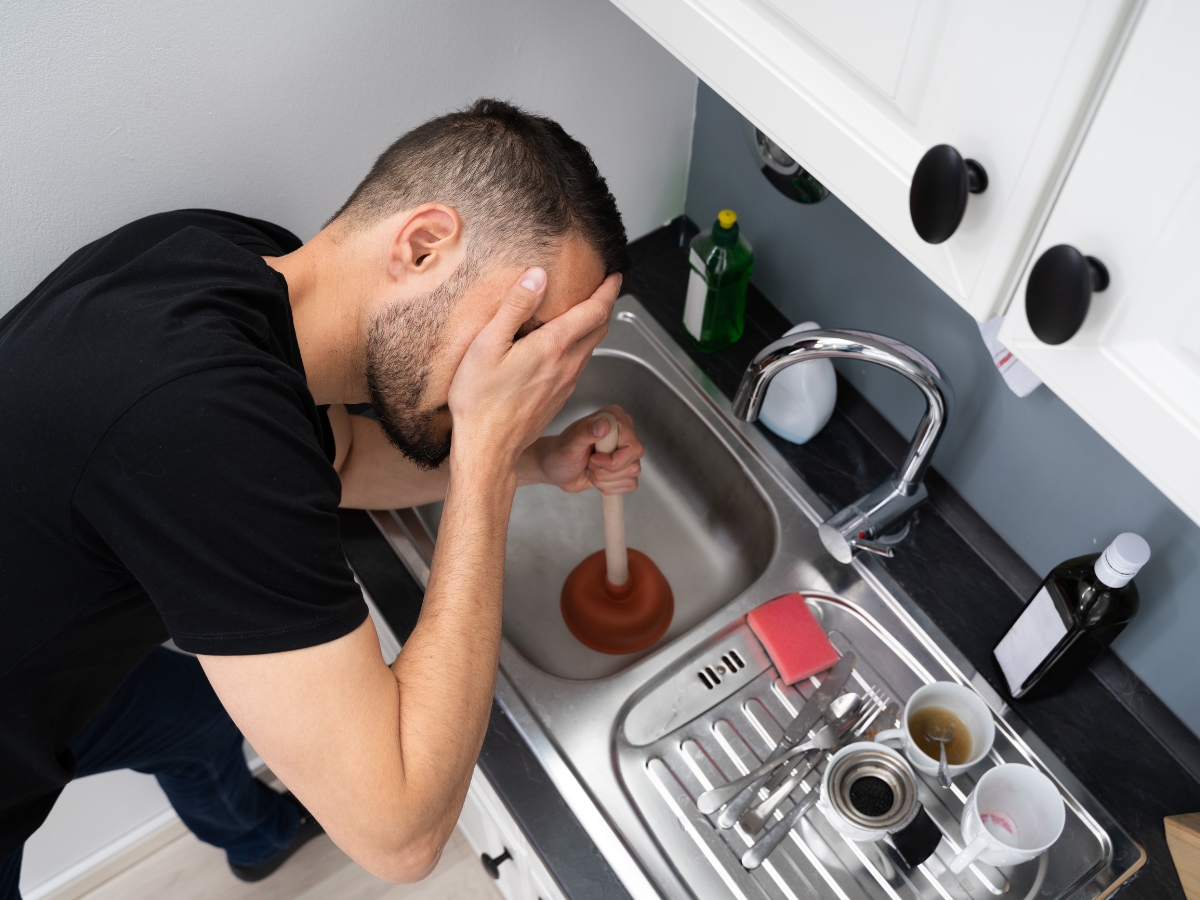 Holiday Septic Tips in Acworth, GA: Preparing for Guests 4 A man holds his face in frustration while using a plunger in a kitchen sink filled with dirty dishes and utensils—reminding everyone to follow Holiday Septic Tips to avoid plumbing headaches this season.