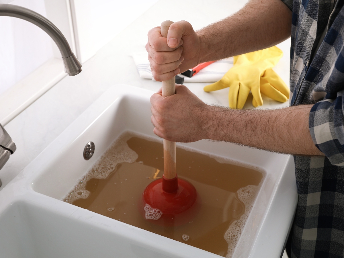Holiday Septic Tips in Acworth, GA: Preparing for Guests 6 A person uses a plunger to unclog a kitchen sink filled with dirty, brownish water; yellow rubber gloves rest nearby on the counter, following Holiday Septic Tips for a cleaner kitchen.