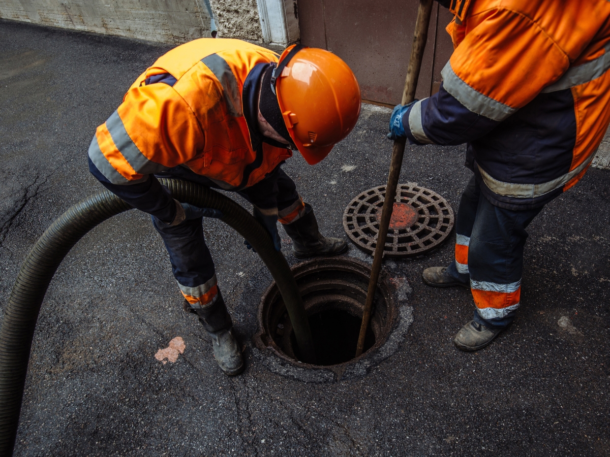 Two workers in orange safety gear clean a manhole with a hose and long tool on an asphalt surface near an open sewer cover, demonstrating important Holiday Septic Tips for safe and efficient maintenance.