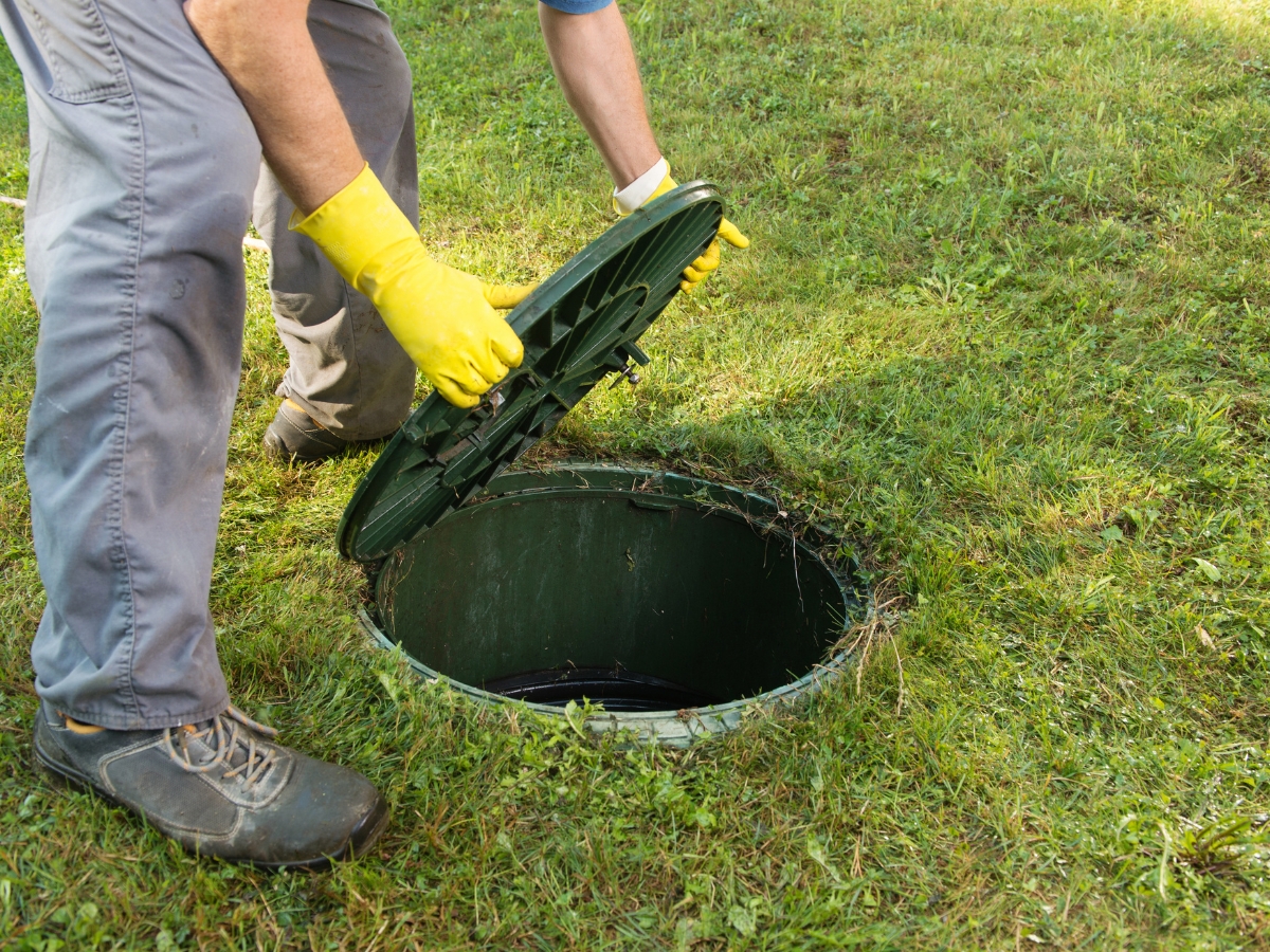 Wearing yellow gloves, a person opens the lid of an outdoor septic tank embedded in the grass, following a year-end septic checklist in Kennesaw.