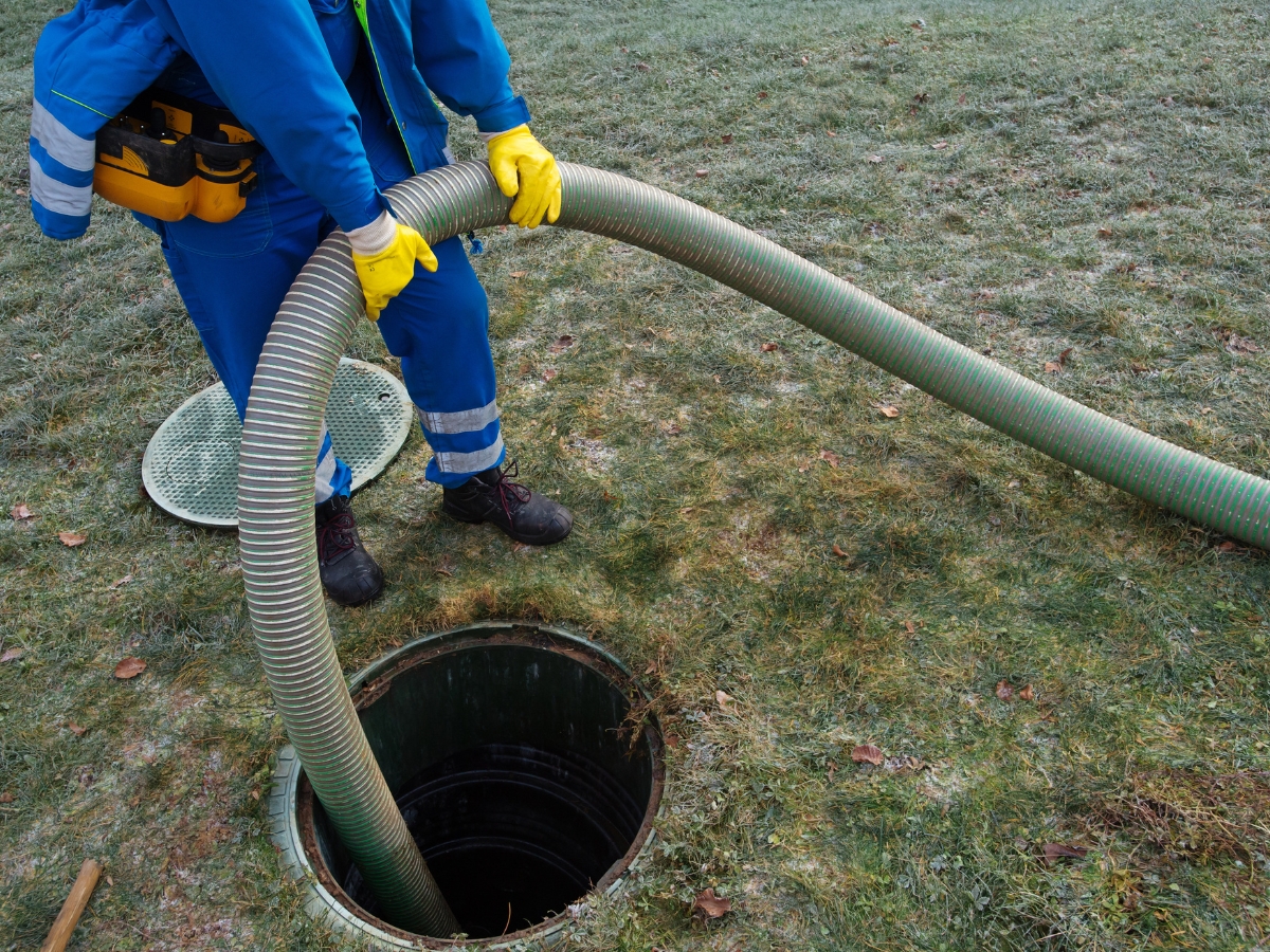 Wearing safety gear, a person uses a large hose to service an open septic tank on grassy ground—essential for effective winter septic system maintenance.