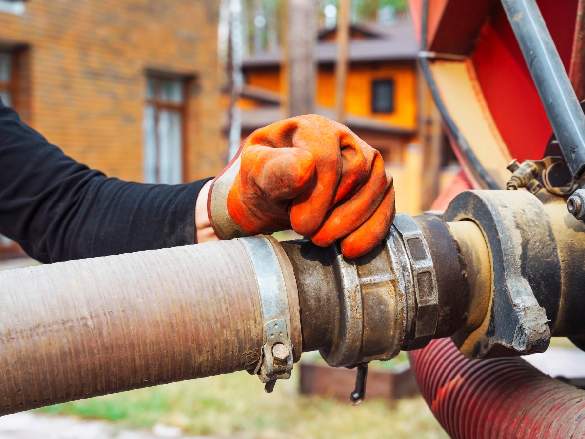 A person wearing orange work gloves is gripping a large hose connected to industrial equipment, demonstrating winter septic system maintenance with buildings visible in the background.