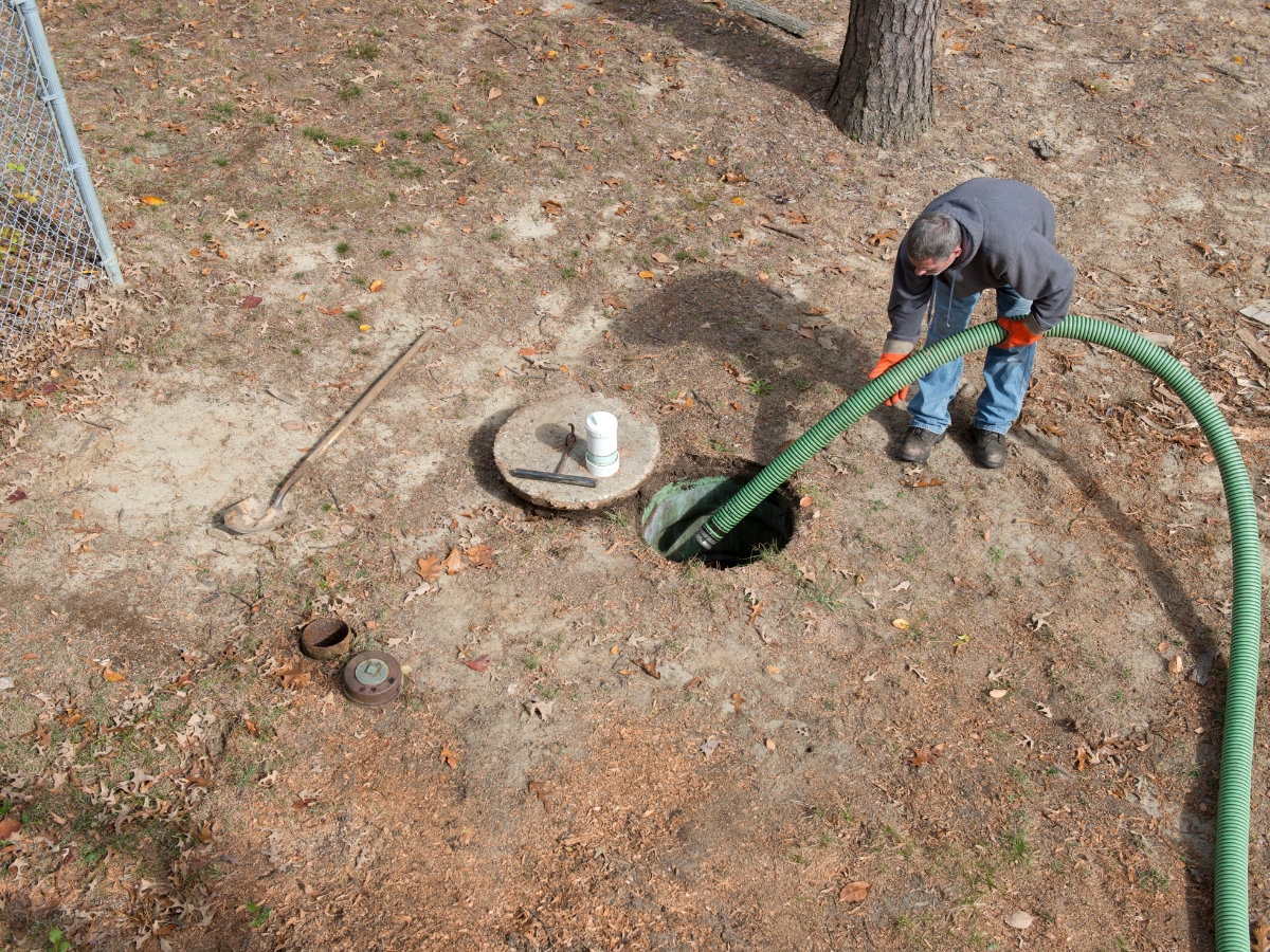A man uses a large green hose to pump out a septic tank in a dirt yard, with septic additives, a shovel, and the septic access cap nearby.